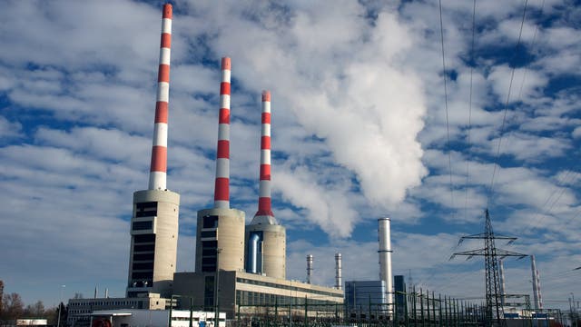 Ein großes Kraftwerk mit mehreren hohen Schornsteinen, die abwechselnd rot-weiß gestreift sind, ragt in den Himmel. Aus einem der Schornsteine steigt weißer Rauch auf. Im Vordergrund sind Stromleitungen und ein verschneites Feld zu sehen. Der Himmel ist teils bewölkt.