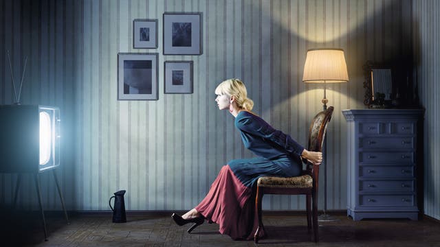 Elegant gekleidete Frau beugt sich in abgedunkeltem Zimmer zu einem eingeschalteten Fernseher hin und hält sich vor Spannung am Stuhl fest.
