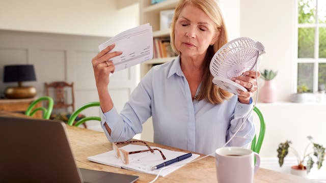 Eine Frau sitzt an einem Holztisch in einem hellen Raum. Sie hält einen kleinen Ventilator in der einen Hand und wedelt mit einem Papier in der anderen Hand, um sich abzukühlen. Vor ihr liegt ein Laptop, eine Brille auf einem Notizblock und eine Tasse Kaffee. Im Hintergrund sind Bücherregale und Pflanzen zu sehen. 