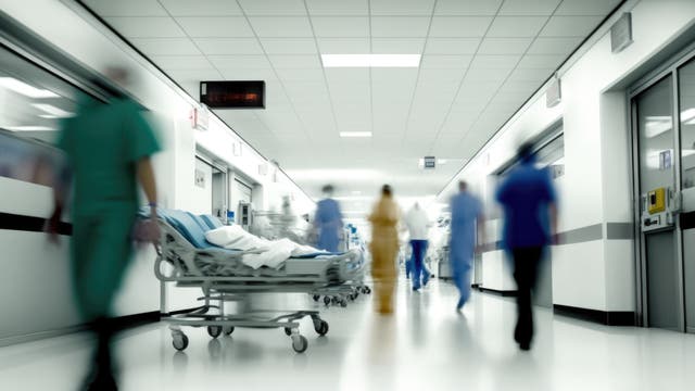 Ein Krankenhausflur mit mehreren Personen in medizinischer Kleidung, die sich bewegen. Im Vordergrund steht ein leeres Krankenhausbett. Die Umgebung ist hell beleuchtet, und an der Decke hängt eine digitale Uhr. Die Szene vermittelt eine geschäftige, aber geordnete Atmosphäre.