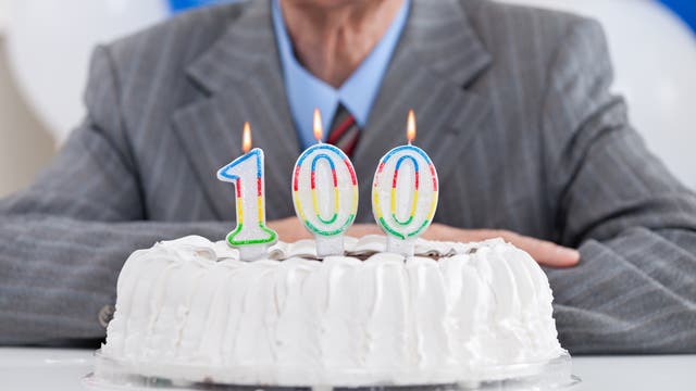 Eine weiße Torte mit drei brennenden Kerzen in Form von den Ziffern 1, 0, 0; im Hintergrund unscharf ein anzugtragender Mann 