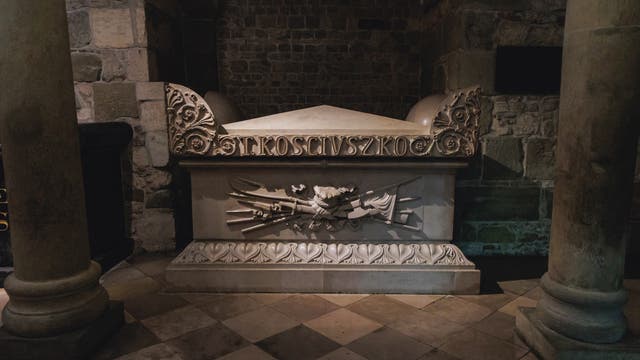 Ein steinernes Grabmal in der Wawel-Kathedrale, umgeben von Säulen und Steinwänden. Auf dem Grabmal ist der Name "KOSCIUSZKO" eingraviert. Verzierungen und Reliefs schmücken die Oberfläche, darunter gekreuzte Fahnen und ein Schild. 