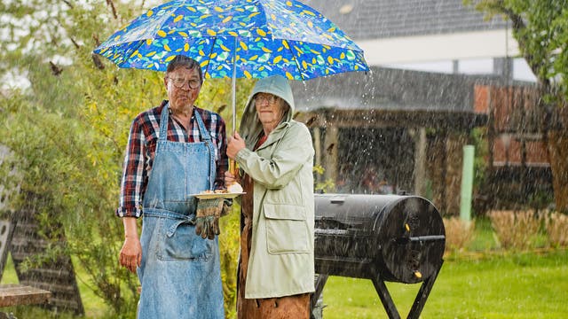 Ein älteres Paar steht im Regen unter einem bunten Regenschirm. Der Mann trägt eine Schürze und hält einen Teller mit Essen, während die Frau einen Regenmantel trägt. Im Hintergrund sind ein Grill und ein Garten zu sehen. 