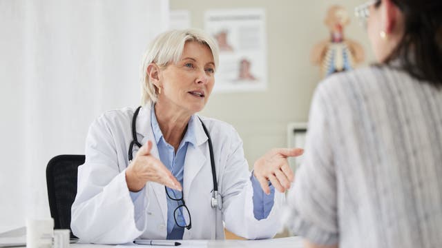 Eine Ärztin mit grauem Haar und Stethoskop spricht mit einer Patientin in einem Behandlungszimmer. Die Ärztin gestikuliert, während sie der Patientin zuhört. Im Hintergrund sind medizinische Diagramme sichtbar.