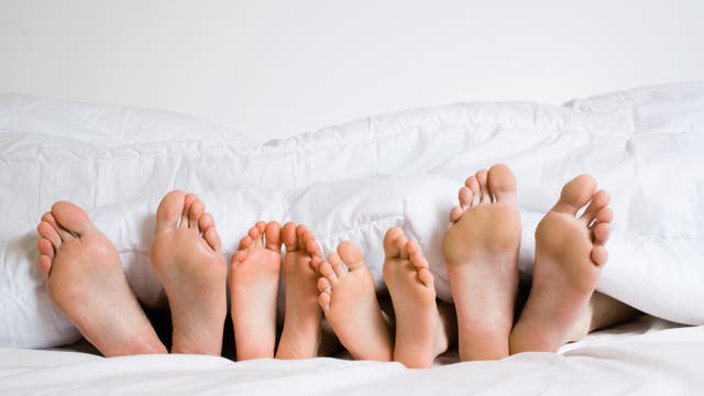 Man sieht die Füße von vier Personen unter einer Bettdecke hervorragen: zwei große Paare, zwei kleine Paare in der Mitte