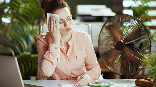 Eine Frau sitzt neben einem Computer und einem Ventilator und hält sich erschöpft ein Tuch an die Stirn