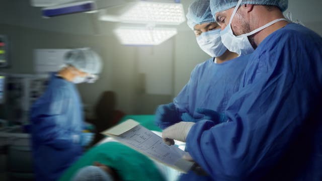 Im Vordergrund studieren zwei Ärzte in OP-Kleidung die Patienten-Werte. Im Hintergrund bereitet jemand die Geräte für die OP vor.
