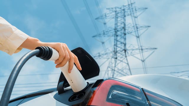 Eine Person lädt ein Elektroauto auf: man sieht ihre Hand, die ein Ladekabel in den Ladeanschluss des Fahrzeugs steckt. Im Hintergrund ist ein Strommast zu sehen, der den Bezug zur Stromversorgung und erneuerbaren Energien herstellt. Der Himmel ist klar und blau.