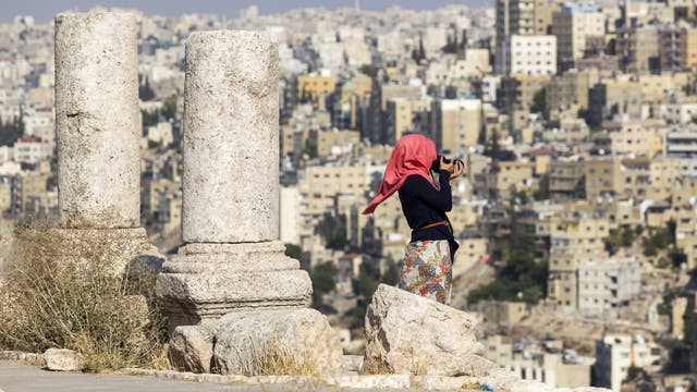 Eine junge Frau mit rotem Kopftuch steht mit einer Kamera filmend neben zwei römischen Säulenstümpfen vor den Häusern von Amman