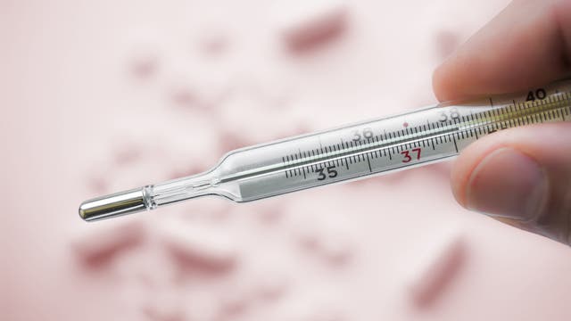 Ein zwischen Daumen und Zeigefinger gehaltenes klassisches Thermometer mit Quecksilber vor rosafarbenem Hintergrund, dort liegen einzelne Tabletten.