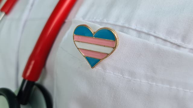 Ein Stethoskop mit roten Schläuchen hängt um den Hals einer Person in einem weißen Kittel. An der Brusttasche des Kittels ist ein herzförmiger Anstecker mit den Farben Blau, Rosa und Weiß befestigt, der die Transgender-Flagge darstellt.