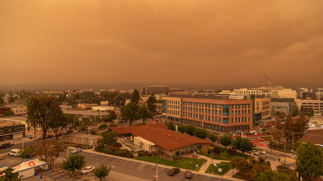 Städtische Landschaft unter einem orangefarbenen Himmel, der durch Rauch von Waldbränden verursacht wird. Im Vordergrund sind Gebäude und ein Parkplatz zu sehen, während im Hintergrund weitere städtische Strukturen und Bäume sichtbar sind. Die Atmosphäre wirkt düster und ungewöhnlich gefärbt.