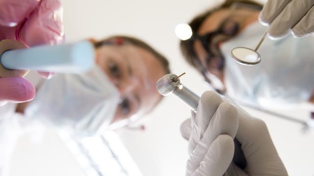 Aus der Perspektive eines Patienten im Zahnarztstuhl: ein Arzt und eine Helferin mit Bohrer und Absauger beugen sich über den Patienten.