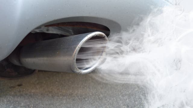 Nahaufnahme eines Autoauspuffs, aus dem weißer Rauch austritt. Der Auspuff ist metallisch und zeigt Anzeichen von Rost. Der Rauch deutet auf laufenden Motorbetrieb hin, möglicherweise bei kaltem Wetter. Das Bild thematisiert Umweltverschmutzung und Fahrzeugemissionen.