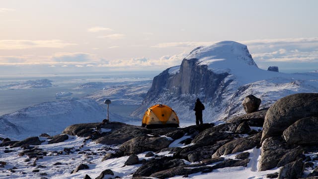 Ein in Schwarz gekleideter Forscher steht neben einem gelben Kuppelzelt und einer Messstation auf einem Berg nahe der Küste Grönlands. Die Landschaft ist mit Schnee und Eis bedeckt