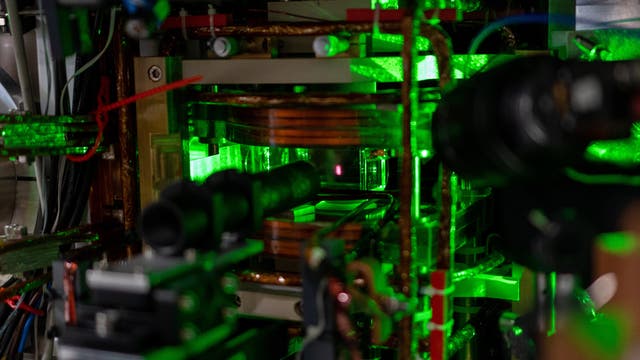 In einer magneto-optischen Falle leuchten inmitten grüner Laserstrahlen und Messaufbauten rötlich Atome, die in der Schwebe gehalten werden