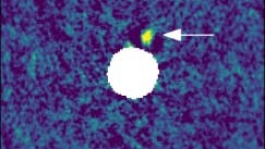 Ein astronomisches Bild zeigt eine farbkodierte Karte des Himmels mit einem zentralen, weißen, kreisförmigen Bereich, der den ausgeblendeten Bereich um den Hauptstern darstellt. Ein Pfeil weist auf einen kleinen, hellen Bereich in der Nähe des Kreises hin, der ein astronomisches Objekt darstellt. Die Achsen sind mit "Δ RA [arcsec]" und "Δ Dec [arcsec]" beschriftet. Eine Skala von "10 au" ist am unteren Rand angegeben.