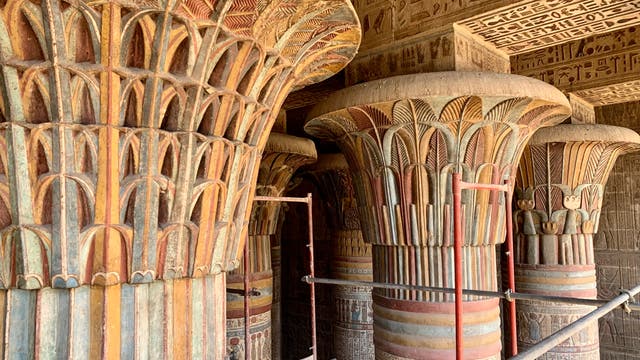 Mehrere reich verzierte, farbenfrohe Säulen im Tempel von Hathor in Dendera, Ägypten. Die Säulen sind mit floralen Mustern und Hieroglyphen verziert. Im Hintergrund sind weitere Säulen und Wände mit antiken ägyptischen Inschriften zu sehen. Ein Gerüst ist zwischen den Säulen aufgebaut, was auf Restaurierungsarbeiten hindeutet.