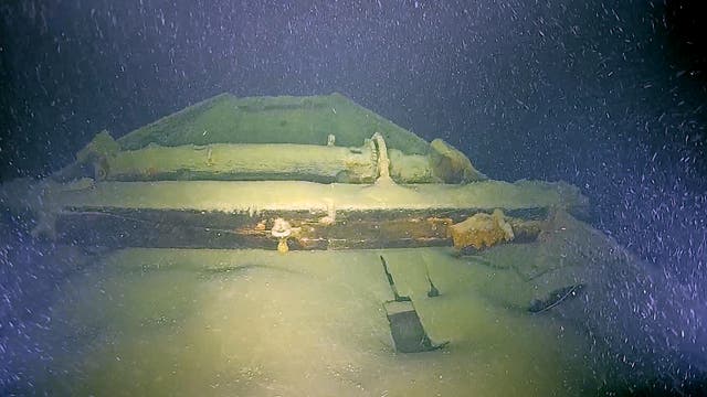 Unterwasseraufnahme eines alten Schiffswracks, das von Sand und Meeresbewuchs bedeckt ist. Die Struktur des Wracks ist teilweise sichtbar, mit erkennbaren Holzbalken und metallischen Elementen. Das Bild zeigt eine ruhige, dunkle Umgebung mit schwebenden Partikeln im Wasser, die die Tiefe und Abgeschiedenheit des Fundorts betonen.