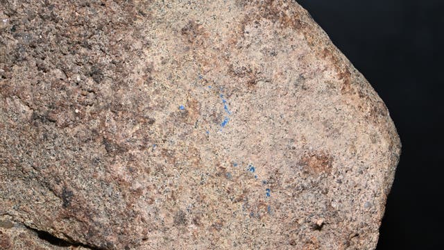 Nahaufnahme eines groben Felsens mit unregelmäßiger Oberfläche und verschiedenen Brauntönen. Der Felsen zeigt kleine blaue Markierungen, die sich über die Oberfläche verteilen. Der Hintergrund ist dunkel und hebt den Felsen hervor.