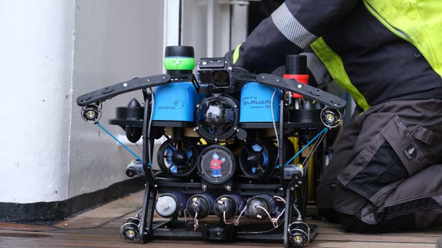Ein Unterwasserroboter mit blauen und schwarzen Komponenten wird von einer Person in gelber Sicherheitskleidung auf einem Holzdeck vorbereitet. Der Roboter trägt die Aufschrift „GEOMAR“ und „Käpt`n Blaubär“. Er ist mit mehreren Kameras und Sensoren ausgestattet. Die Szene findet in der Nähe einer Tür statt, die zu einem Innenraum führt.