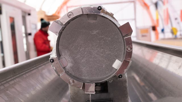 Ein Eisbohrkern befindet sich in einem Rohr des Bohrers, mit dem er aus dem Eis gezogen wurde. Im unscharfen Hintergrund steht ein Forscher mit einem roten Anorak und einer Bommelmütze. Unterhalb des Bohrkerns befindet sich eine Metallwanne