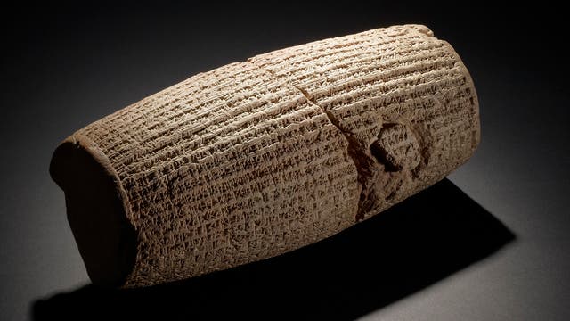 Eine antike, zylindrische Tonrolle mit eingravierten Keilschriftzeichen, die auf einem dunklen Hintergrund liegt. Die Rolle ist leicht beschädigt, was auf ihr Alter hinweist. Die Beleuchtung hebt die Textur und die Details der Inschriften hervor.