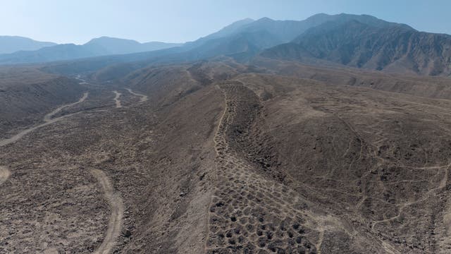Luftaufnahme einer trockenen, hügeligen Landschaft mit erodierten Bergrücken und Tälern. Im Hintergrund sind grüne landwirtschaftliche Felder und ein Flussbett zu sehen, die die karge Wüstenlandschaft kontrastieren. Die Szene zeigt eine lange Reihe von künstlich angelegten Gruben.