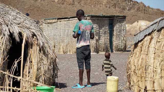 Ein Mann und ein Junge stehen inmitten von Strohhütten auf steinigem Boden.