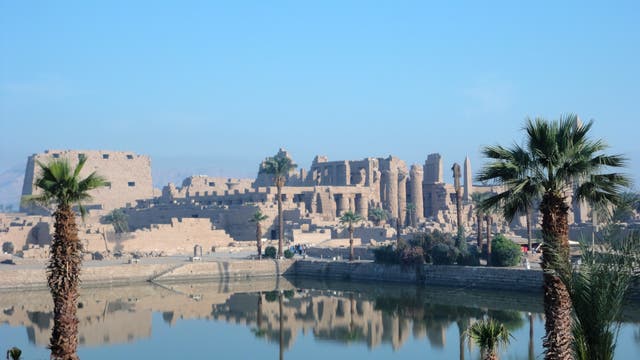 Eine antike Tempelanlage mit massiven Steinstrukturen und Säulen, umgeben von Palmen. Im Vordergrund spiegelt sich die Anlage in einem ruhigen Wasserbecken. Der Himmel ist klar und blau, was die majestätische Atmosphäre der historischen Stätte unterstreicht.