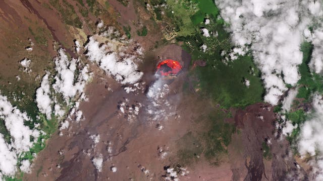Satellitenbild eines Vulkans mit einem aktiven Lavasee in der Mitte. Die Umgebung zeigt eine Mischung aus grüner Vegetation und braunen, vulkanischen Landschaften. Weiße Wolken sind über das Bild verteilt, und die Küstenlinie ist am unteren Rand sichtbar. Das Bild veranschaulicht die geologische Aktivität und die natürliche Umgebung des Vulkans.