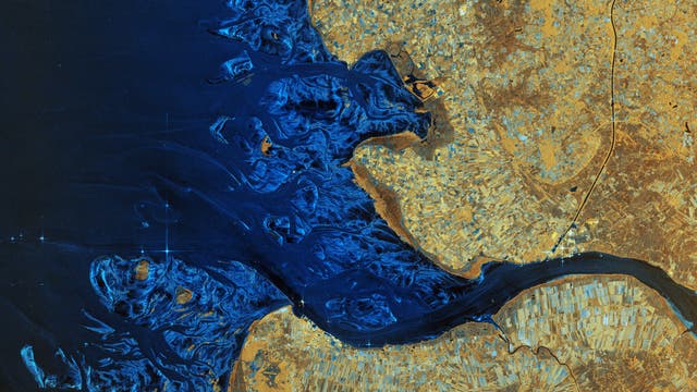 Satellitenbild einer Küstenregion mit dem Flussdelta der Elbe, die in ein tiefblaues Meer mündet. Die linke Bildhälfte zeigt das Meer mit verzweigten Wasserwegen, während die rechte Bildhälfte landwirtschaftlich genutzte Flächen in verschiedenen Brauntönen darstellt. Die Karte hebt die Kontraste zwischen Wasser und Land hervor und zeigt die natürliche Geografie der Region.