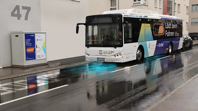 Ein Elektrobus fährt auf einer nassen Straße vor einem Gebäude mit der Nummer 47. Der Bus ist mit der Aufschrift "Laden beim Fahren" versehen und nutzt eine induktive Ladetechnologie, die durch blaue Markierungen auf der Straße angezeigt wird. Links im Bild befindet sich ein Schaltkasten mit der Aufschrift "Laden beim Fahren" und einem QR-Code. Der Bus gehört zum Projekt "e-GELU" und ist mit Logos verschiedener Partner versehen. Im Hintergrund sind moderne Wohngebäude zu sehen.