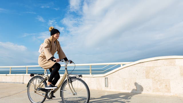 Eine Frau in einem braunen Mantel fährt an einer Strandpromenade Fahrrad.