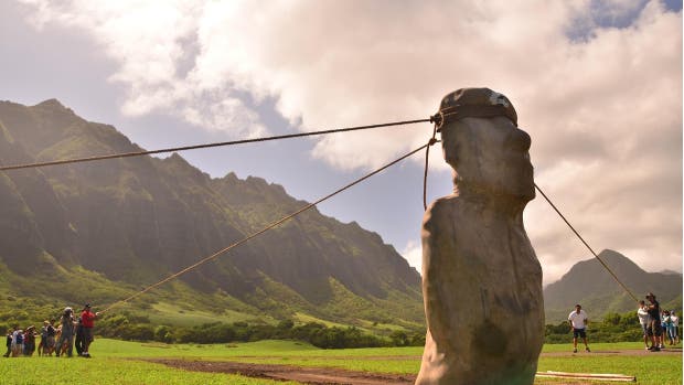 Eine Gruppe von Menschen zieht an Seilen, die an einer großen Statue befestigt sind, um sie aufrecht zu halten. Die Szene spielt sich auf einer grünen Wiese vor einer beeindruckenden Bergkulisse ab. Der Himmel ist teilweise bewölkt, und die Sonne scheint. Die Menschen scheinen an einer Demonstration oder einem Experiment teilzunehmen, das möglicherweise die Bewegung oder Stabilität der Statue untersucht.