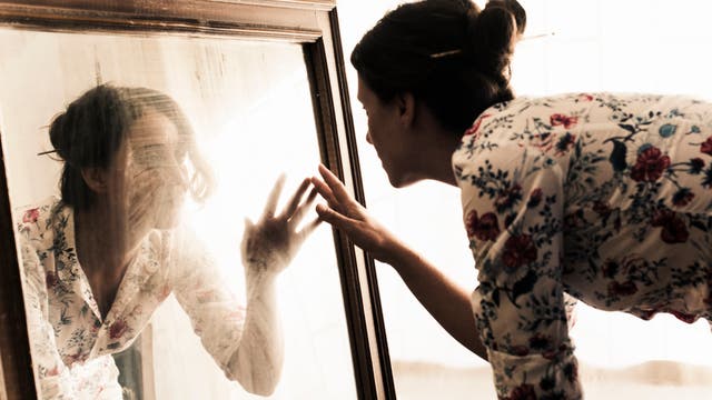 Eine Frau kniet vor einem Spiegel und streckt die Hand nach ihrem Spiegelbild aus.