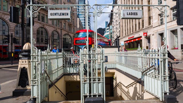 Eine Straßenszene in London mit einem roten Doppeldeckerbus im Hintergrund. Im Vordergrund befindet sich ein Eingang zu einer öffentlichen Toilette mit den Schildern "Gentlemen" und "Ladies". Menschen fahren mit dem Fahrrad oder gehen vorbei. Die Umgebung ist von historischen Gebäuden und modernen Hochhäusern geprägt.