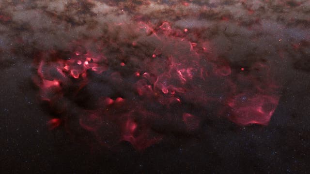Eine astronomische Aufnahme zeigt eine ausgedehnte, rötlich leuchtende Gaswolke im Weltraum, umgeben von dunklen Staubwolken und einem Hintergrund aus zahlreichen kleinen Sternen. Die roten Bereiche deuten auf ionisiertes Wasserstoffgas hin, das durch nahegelegene Sterne zum Leuchten angeregt wird. Die Struktur der Wolke ist unregelmäßig und weist verschiedene Dichtebereiche auf, die durch die dunklen und hellen Kontraste sichtbar werden.