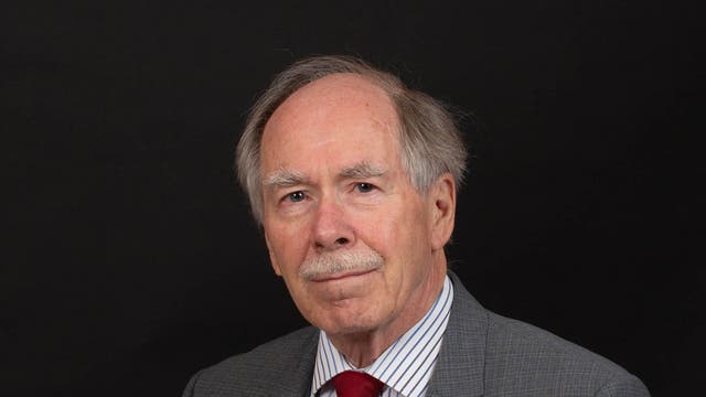 Gerard 't Hooft, ein älterer Mann mit Schnurrbart in einem grauen Anzug, blickt vor einem dunklen Hintergrund in die Kamera.