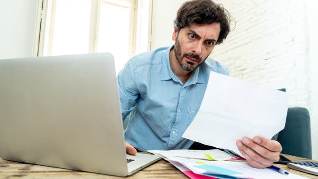 Ein Mann in einem hellblauen Jeanshemd sitzt an einem Holztisch und schaut mit weit aufgerissenen Augen auf ein Dokument in seiner Hand. Vor ihm steht ein Laptop, und auf dem Tisch liegen weitere Papiere. Im Hintergrund sind ein Fenster und eine weiße Backsteinwand zu sehen.