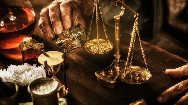 Eine Hand hält ein Glas mit goldenen Nuggets und gießt sie auf eine antike Waage auf einem Holztisch. Neben der Waage stehen eine Sanduhr, ein Glas mit roter Flüssigkeit und Kristalle. Die Szene erinnert an alchemistische Praktiken.
