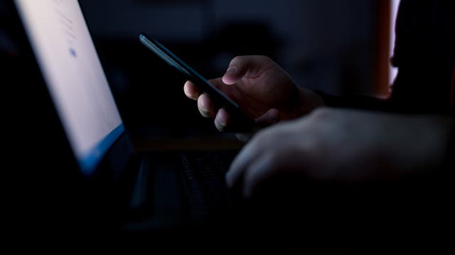 Eine Person sitzt in einem dunklen Raum vor einem Laptop und hält ein Smartphone in der Hand. Der Bildschirm des Laptops leuchtet schwach und beleuchtet die Hände der Person. Die Szene vermittelt eine Atmosphäre von Konzentration und Technologie.