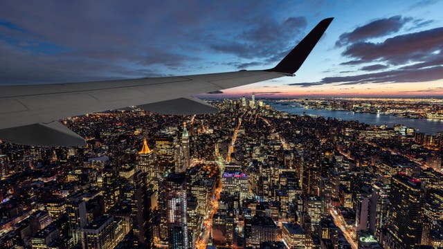 Blick aus einem Flugzeugfenster auf eine beleuchtete Stadtlandschaft bei Sonnenuntergang. Die Flügelspitze des Flugzeugs ist im Vordergrund sichtbar, während sich darunter ein dichtes Netz aus erleuchteten Straßen und Gebäuden erstreckt. Der Himmel zeigt eine Mischung aus Blau- und Orangetönen, und ein Fluss ist am Horizont zu erkennen.