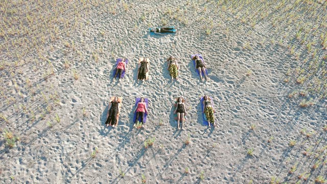 Eine Gruppe von Menschen liegt auf dem Rücken auf einem sandigen Boden, verteilt in zwei Reihen. Sie sind auf bunten Matten positioniert, umgeben von spärlichem Pflanzenwuchs. Die Szene wirkt ruhig und entspannt, möglicherweise Teil einer Yoga- oder Meditationsübung im Freien. Die Schatten der Personen sind lang und deuten auf eine tief stehende Sonne hin.