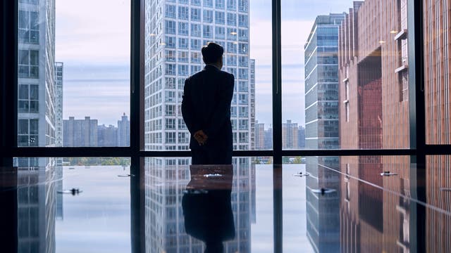 Eine Person in formeller Kleidung steht mit dem Rücken zur Kamera und blickt aus einem großen Fenster auf eine städtische Skyline mit modernen Hochhäusern. Die Reflexion der Person und der Gebäude ist auf einem glänzenden Tisch sichtbar. Die Szene vermittelt einen Eindruck von Nachdenklichkeit und urbaner Geschäftigkeit.