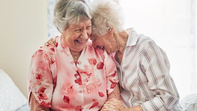 Zwei ältere Frauen sitzen nebeneinander und lachen herzlich. Die Frau links trägt eine rosa Bluse mit Blumenmuster, während die Frau rechts eine gestreifte Bluse trägt. Sie scheinen eine enge Freundschaft zu pflegen, da die Frau rechts ihren Kopf auf die Schulter der anderen lehnt. Die Szene vermittelt Wärme und Freude.