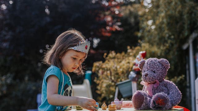 Ein Kind spielt im Freien an einem Tisch mit einem Teeservice und einem lila Teddybär. Das Kind trägt eine Krone und eine Halskette. Im Hintergrund sind Bäume und ein unscharfer Garten zu sehen. Die Szene vermittelt eine spielerische und fantasievolle Atmosphäre.