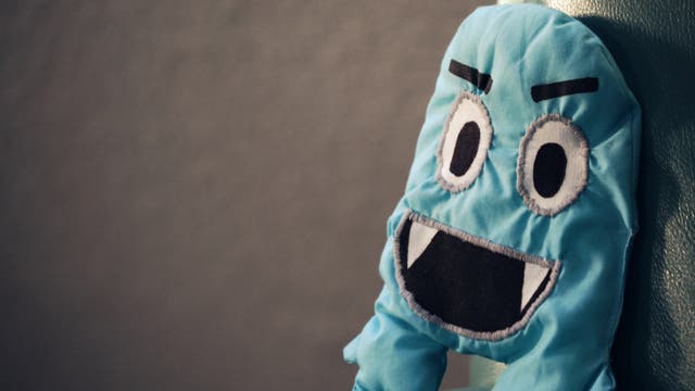 Ein blaues Stoffspielzeug mit großen, aufgenähten Augen und einem breiten Mund sitzt auf einem grünen Ledersofa. Das Spielzeug hat eine cartoonartige Erscheinung mit aufgestickten Augenbrauen und Zähnen. Der Hintergrund ist unscharf und dunkel, was den Fokus auf das Spielzeug lenkt.