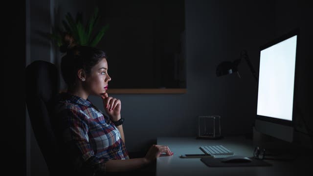 Eine Frau sitzt in einem dunklen Raum vor einem beleuchteten Computerbildschirm. Sie trägt ein kariertes Hemd und stützt ihr Kinn auf ihre Hand, während sie konzentriert auf den Bildschirm schaut. Auf dem Schreibtisch vor ihr liegen eine Tastatur, eine Maus, ein Smartphone und eine Brille.