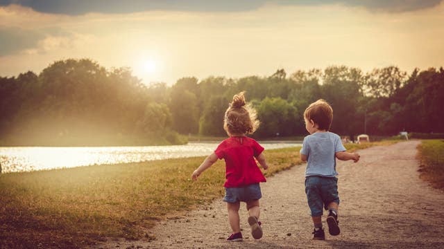 Zwei kleine Kinder laufen auf einem Schotterweg an einem Fluss in Richtung Sonnenuntergang. Das Mädchen trägt ein rotes und der Junge ein hellblaues T-Shirt. Der Himmel ist teils bewölkt und die Sonne steht tief am Horizont. Im Hintergrund sind Bäume und eine ruhige Landschaft zu sehen. Die Szene vermittelt ein Gefühl von Freiheit und Unbeschwertheit.